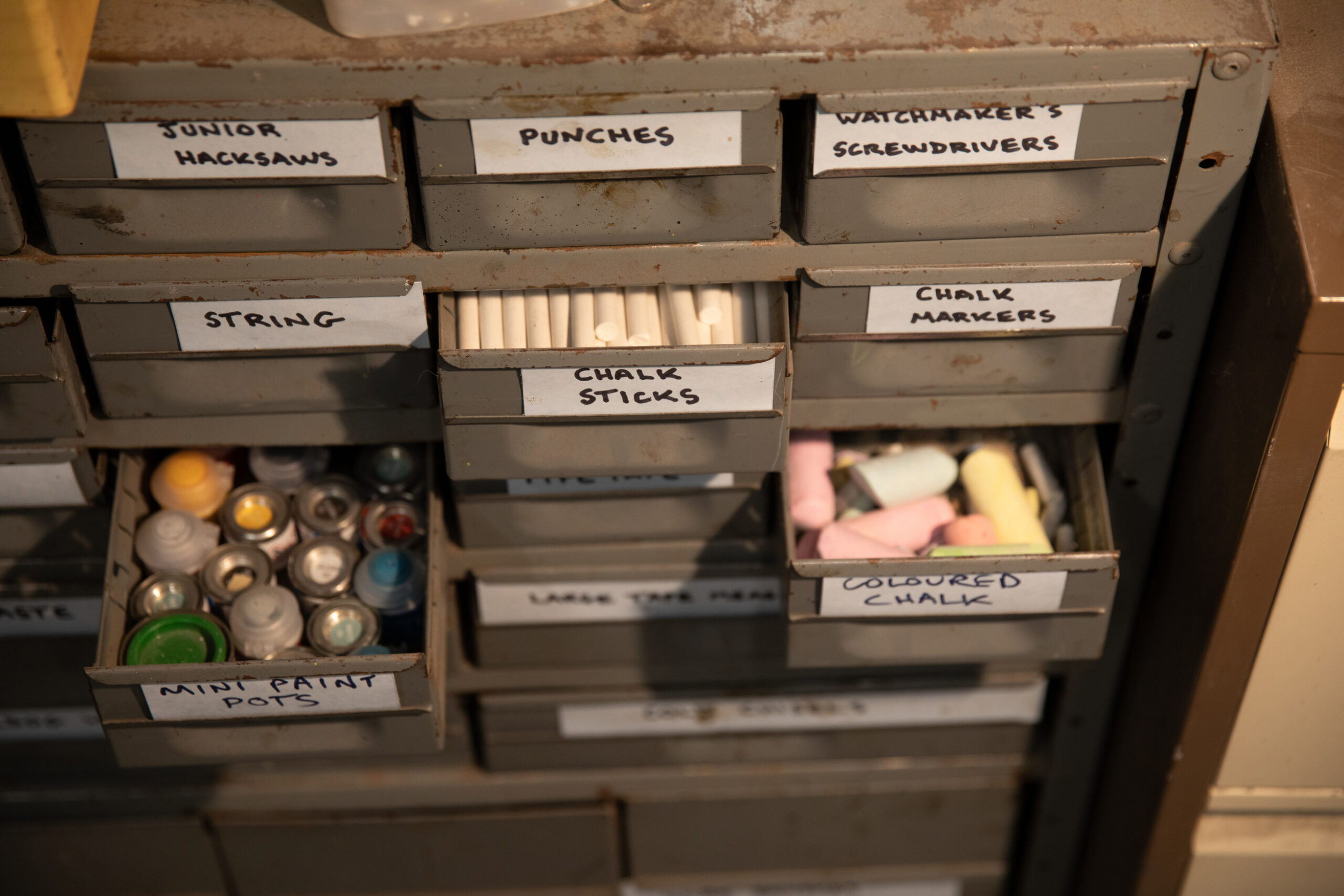 A bank of small drawers with hand-written labels. Some are half-open, showing coloured chalk and small paint pots