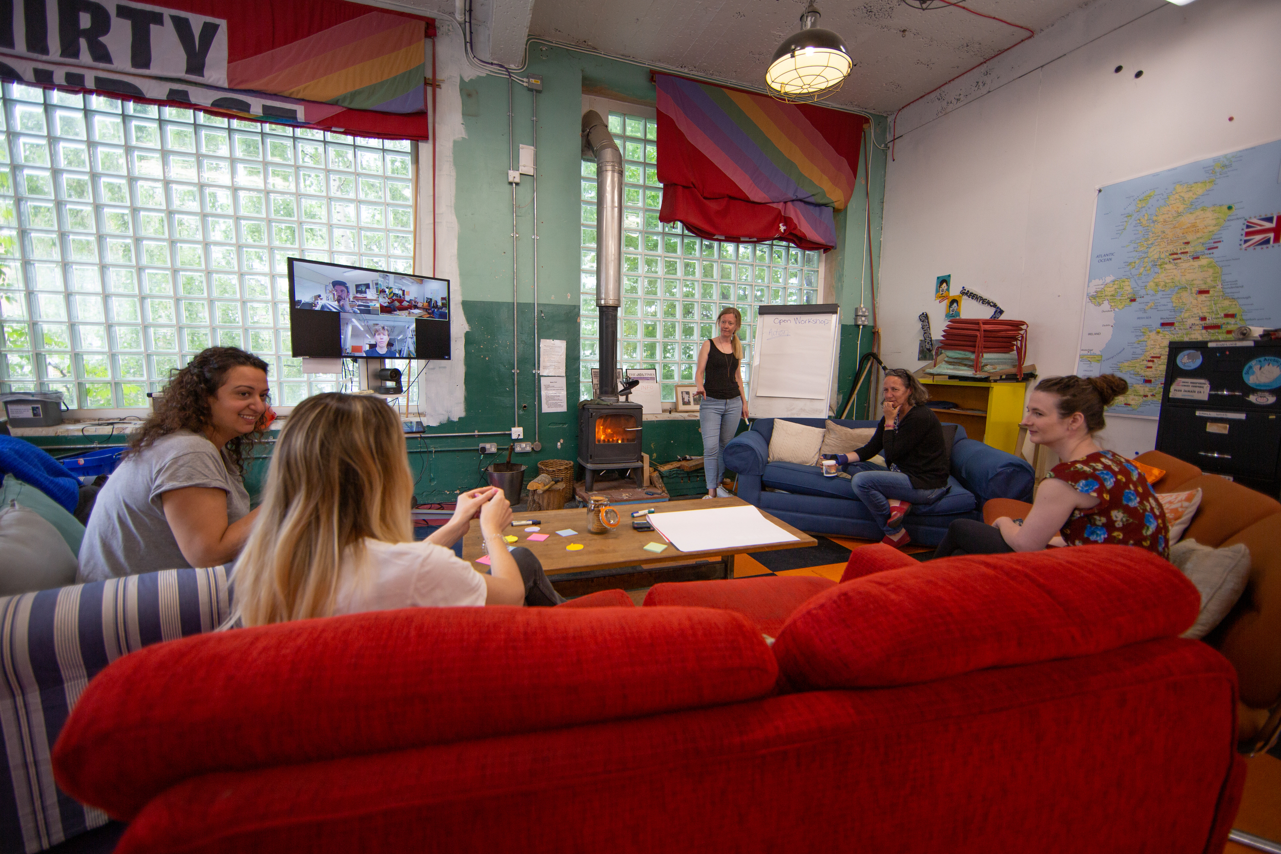 People sitting on sofas gathered around a wood burning stove in a high-ceilinged workshop space.