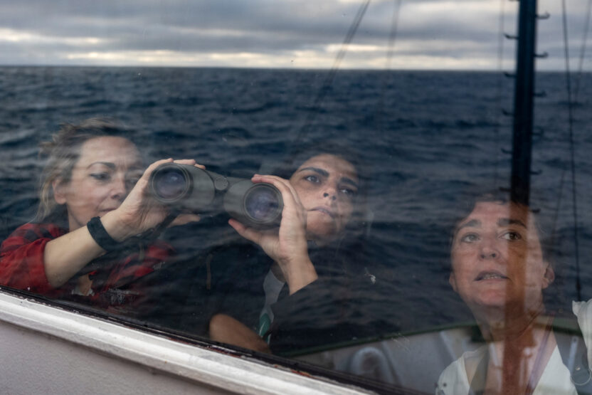 Three crew members look through binoculars out of a window. The ocean and evening sky are reflected in the window.