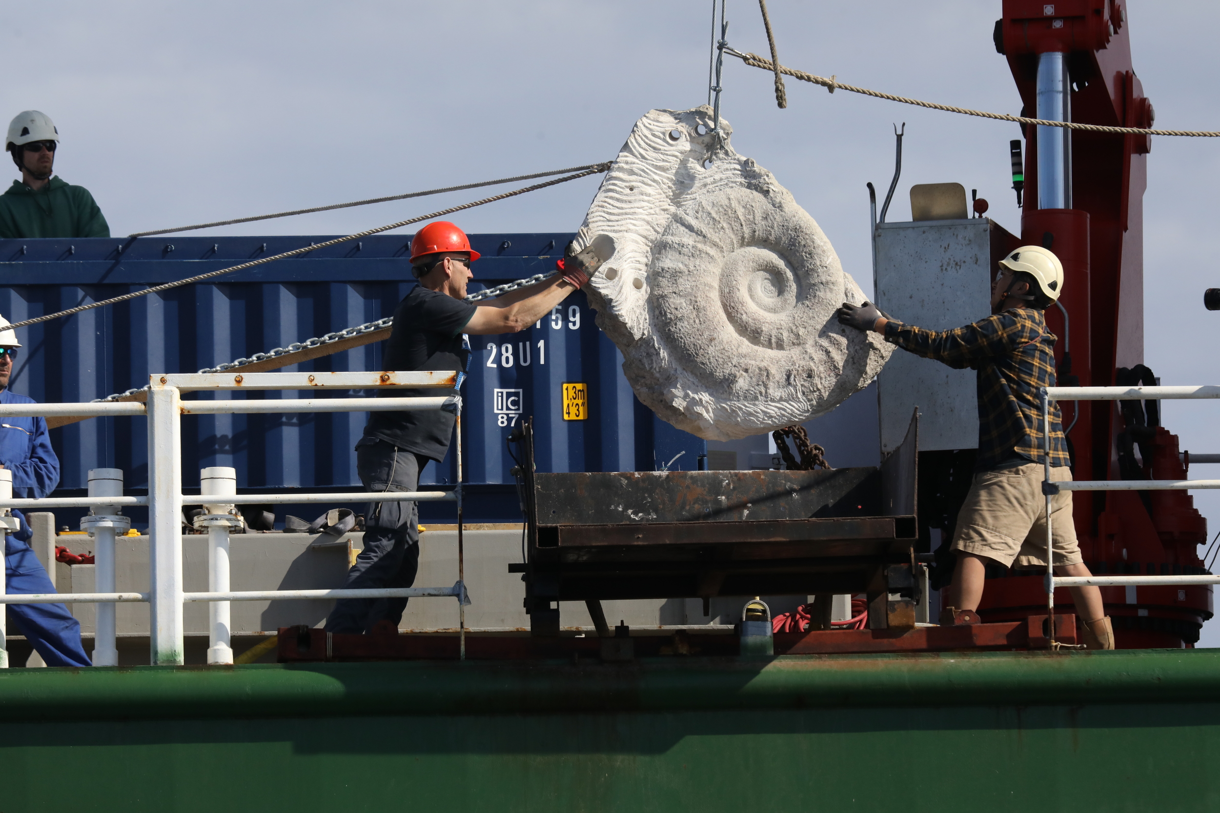 Two people in hardhats move a large stone boulder, with a shell shape carved into the side, using ropes and pulleys on the side of a ship.