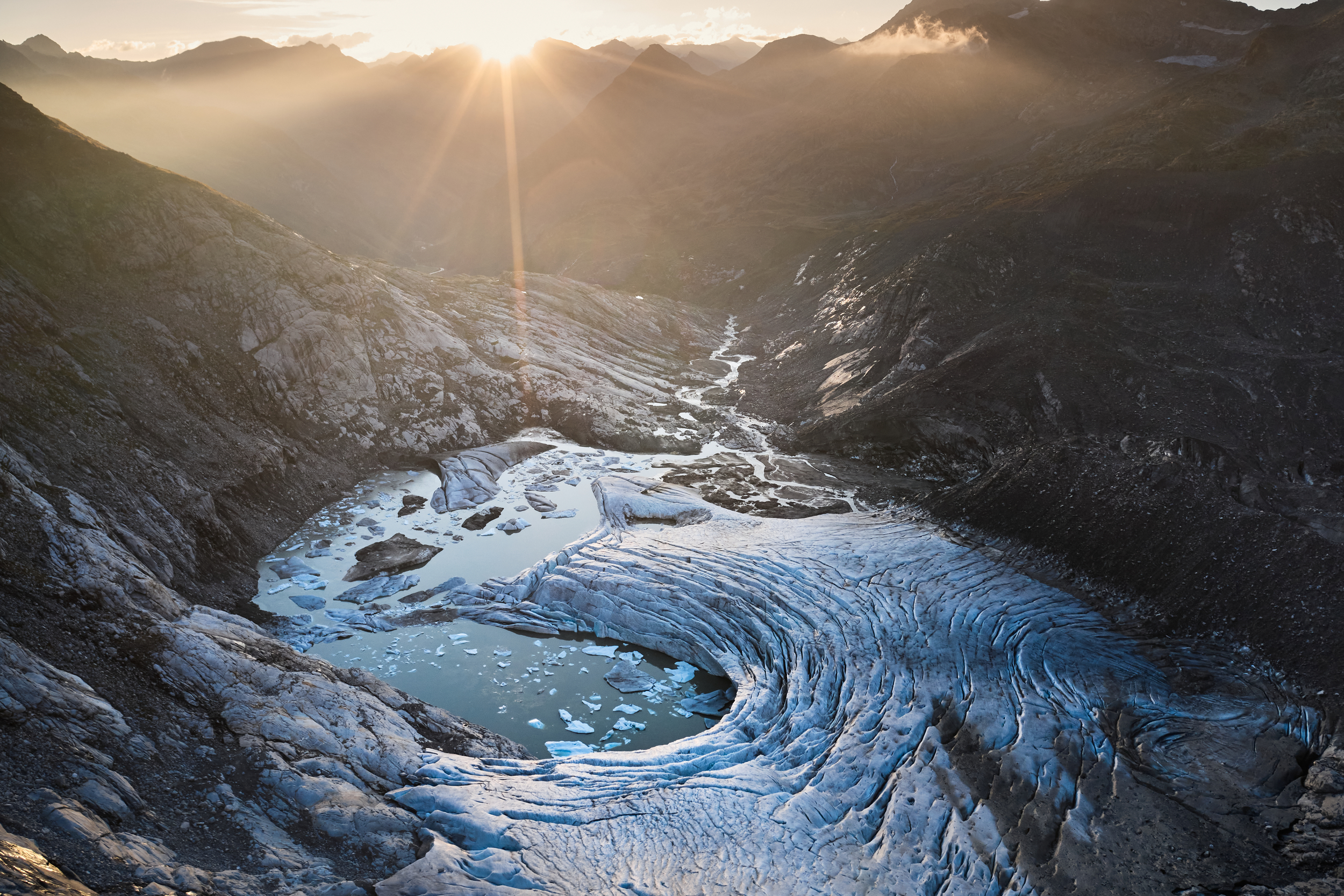 A large swirl of ice with a pool of water in the middle and chips of ice disappearing to a stream in the distance, among rocky mountains, with a sunset peeking just over the top of them.