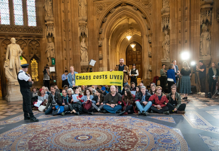 A large group of activists sit in an ornate hall in the Palace of Westminster holding a banner saying 'chaos costs lives'