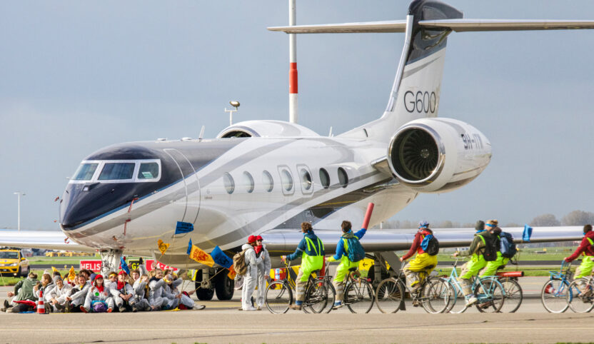 Activists sit around the wheels of a private jet sitting on a runway. More activists on bikes are riding towards them.