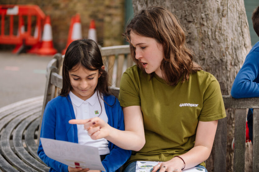 A volunteer wearing a Greenpeace t-shirt sits on a bench outside with a primary school pupil. The volunteer is speaking and pointing to a sheet of paper held by the pupil.