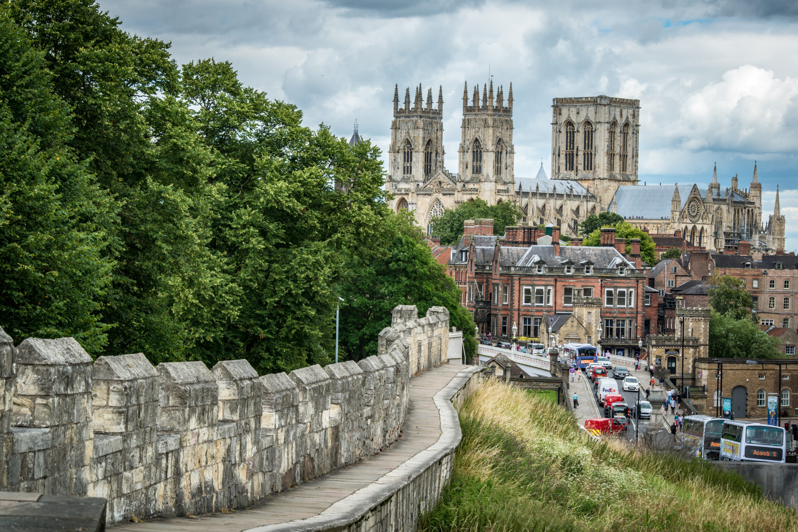 A view of York Minster, with a city wall and a street packed with cars, vans and buses in the foreground.