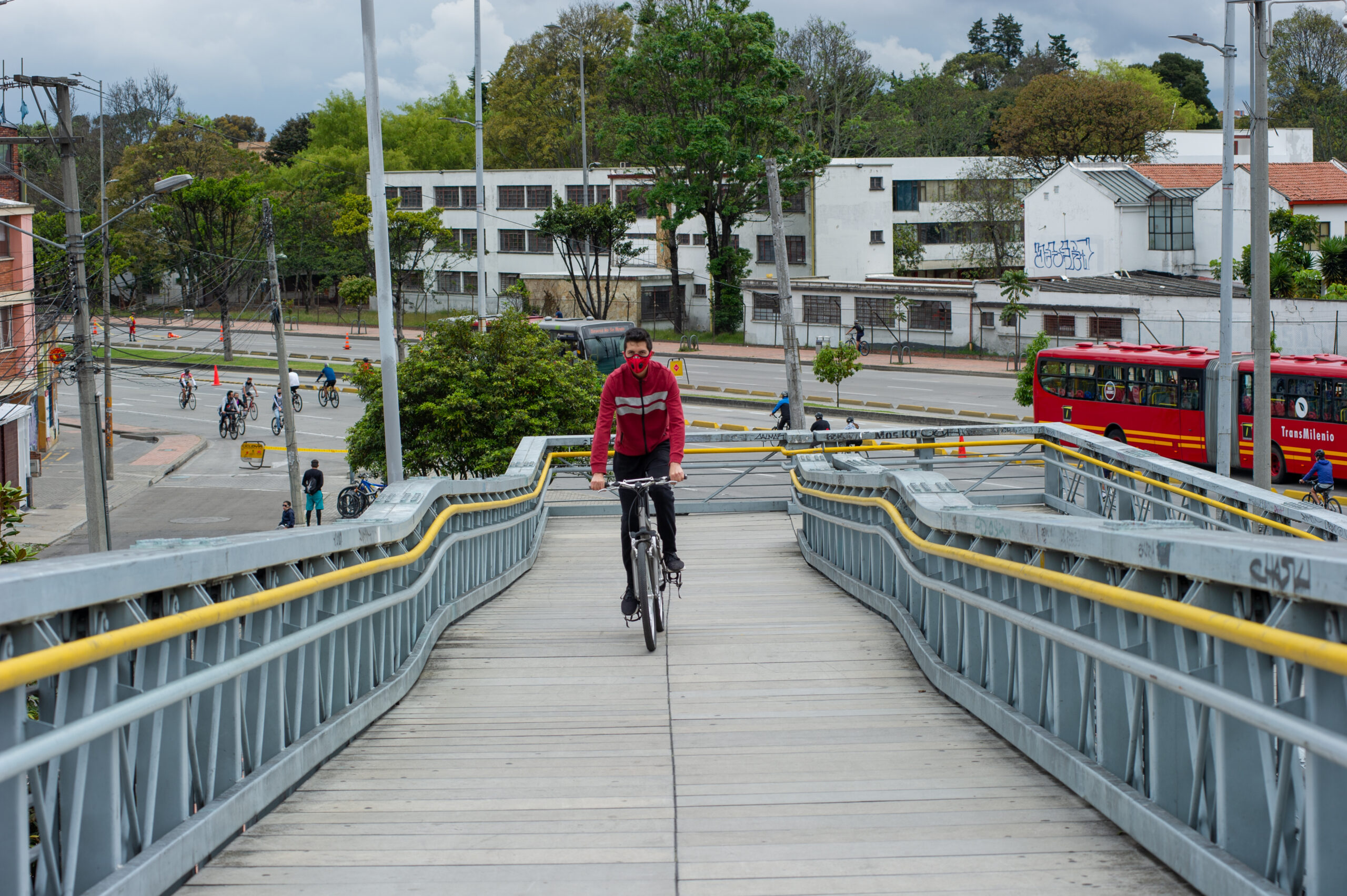 Cyclist climbing a wide bridge designed for cyclists and pedestrians with two levels of handrails and a clear street in the background with a few cyclists and one bus.