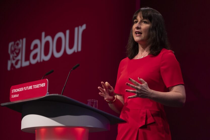 A woman with dark hair in a red dress stands giving a speech in front of a large 