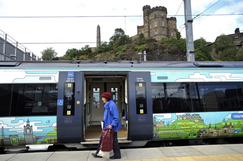 An older woman carrying chopping bags walks past a shiny new train with a castle in the background