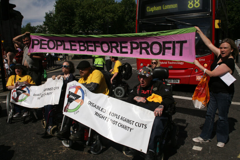 Group of disabled activists protest in a London street. They’re holding banners saying “People before profit” and “Disabled People Against Cuts. Rights not charity”