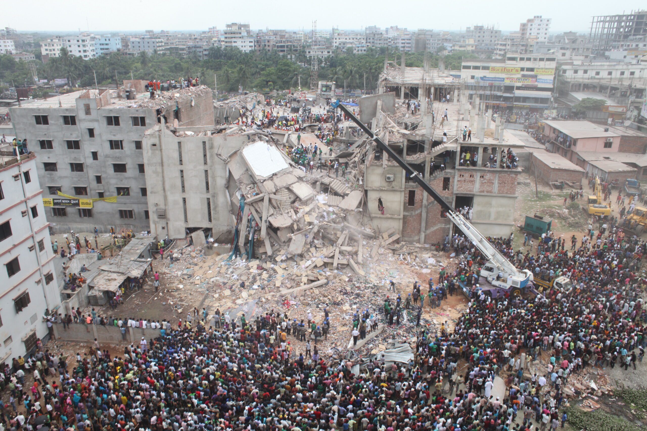 A large crowd around a collapsed factory where rescue workers attempt to save people trapped in the rubble.
