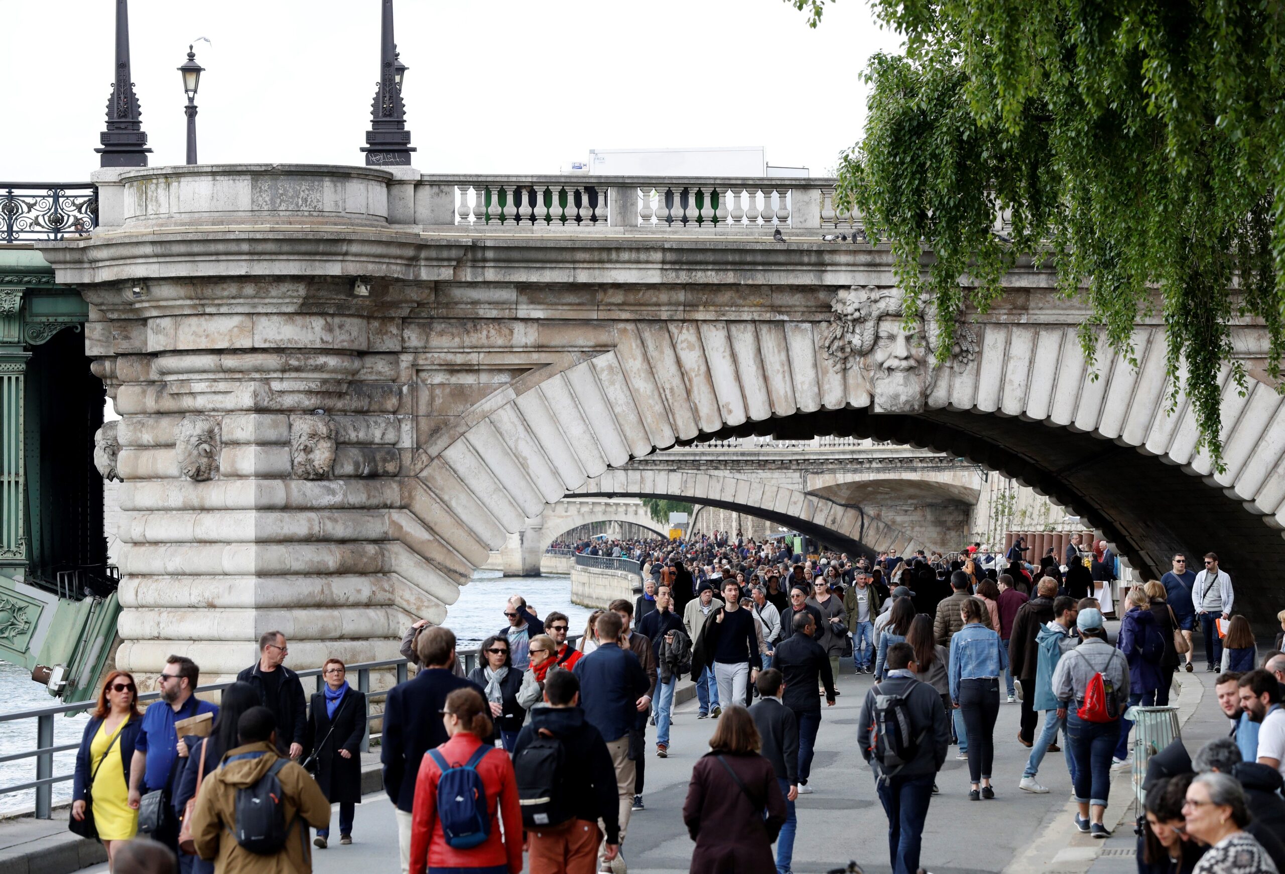 Many people walking along the bank of the River Seine in Paris, on a wide road with the stone arches of multiple bridges over the river in the foreground and distance.