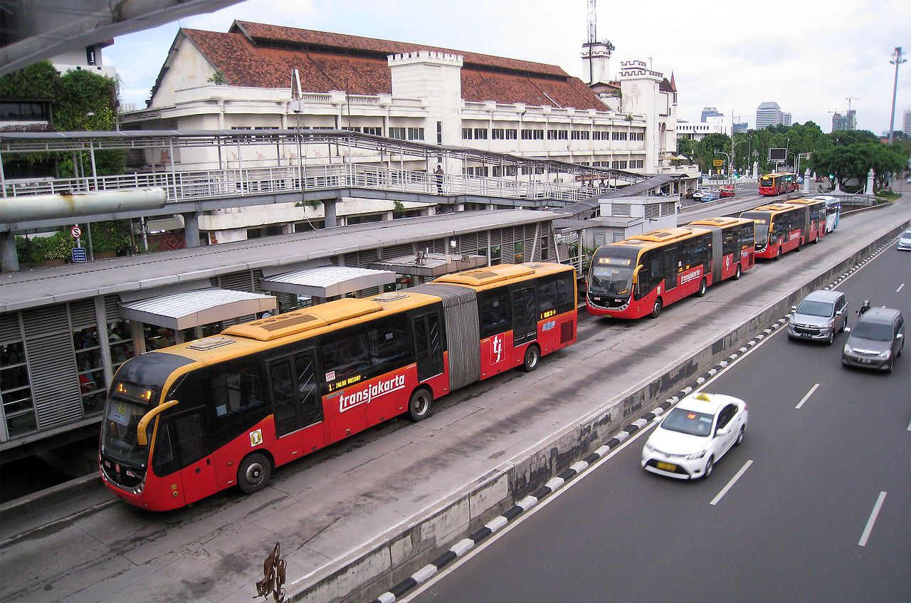 Three red and yellow articulated buses in a separated two lane area stopped to pick up passengers outside a bus terminal featuring multiple buildings and pedestrian bridges, next to a clear road with only four cars.
