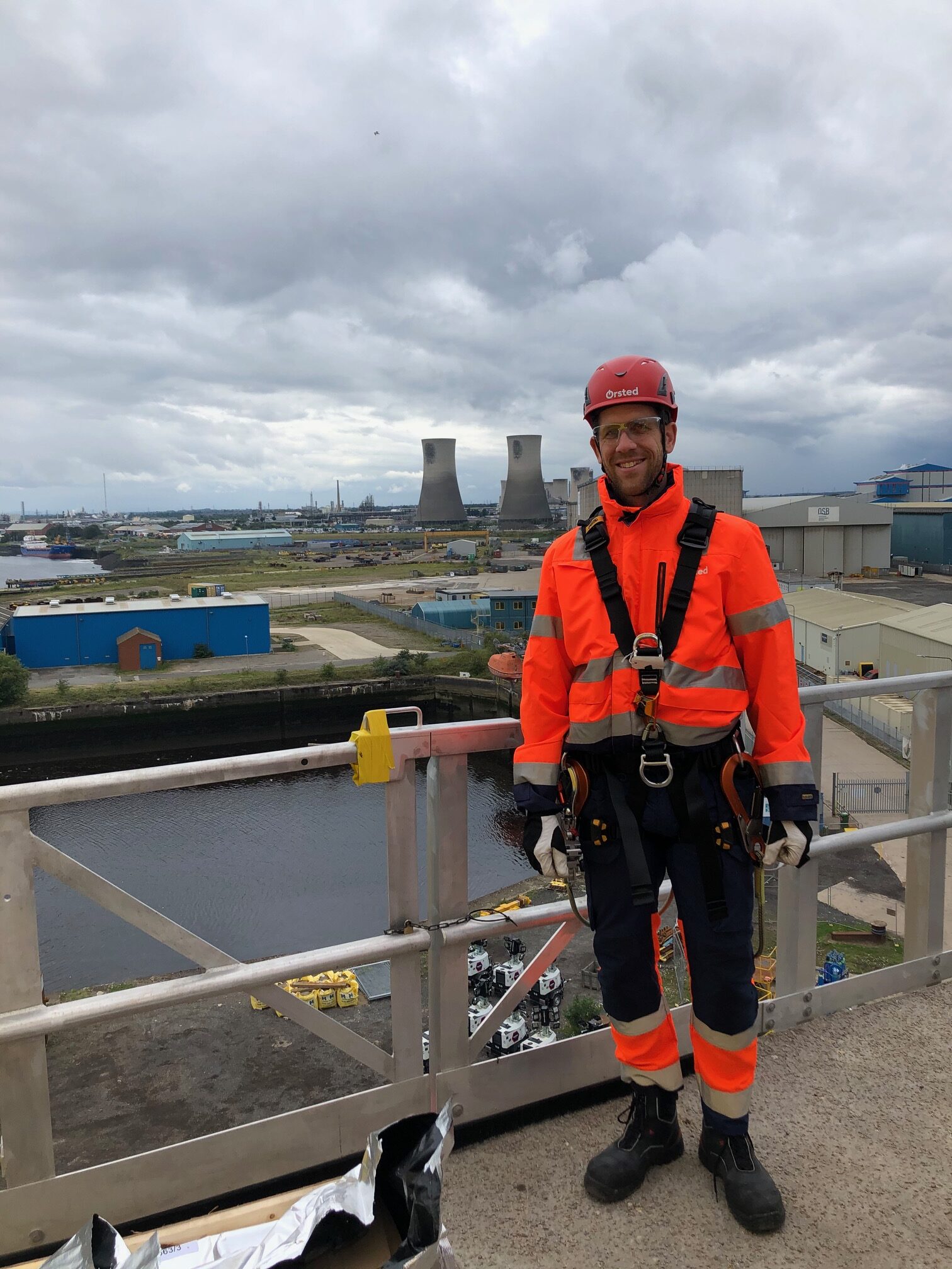 a man standing on a high-up platform with climbing gear on and an industrial zone in the background.