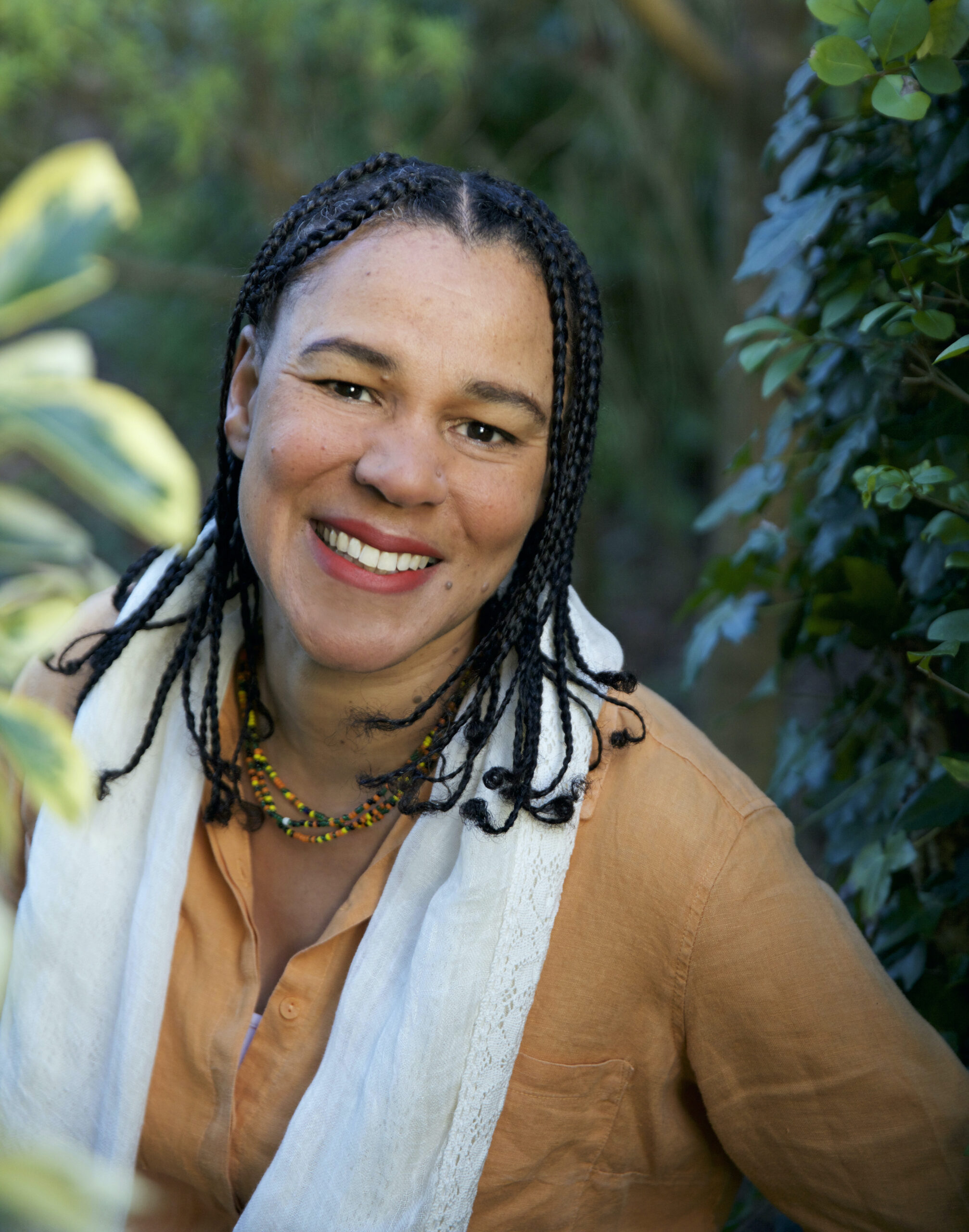 A portrait of a woman with braided hair surrounded by plants smiling.