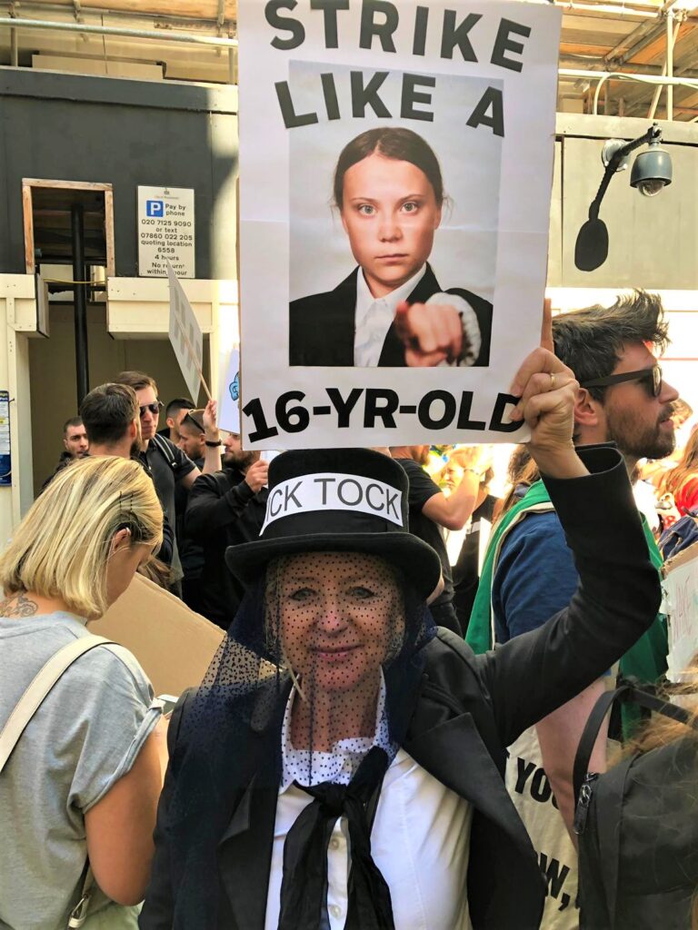 Photograph of Greenpeace volunteer Nicole dressed in mourning at the London Climate Strike, with a sign reading "Strike like a 16 year-old" with around an image of Greta Thunberg.