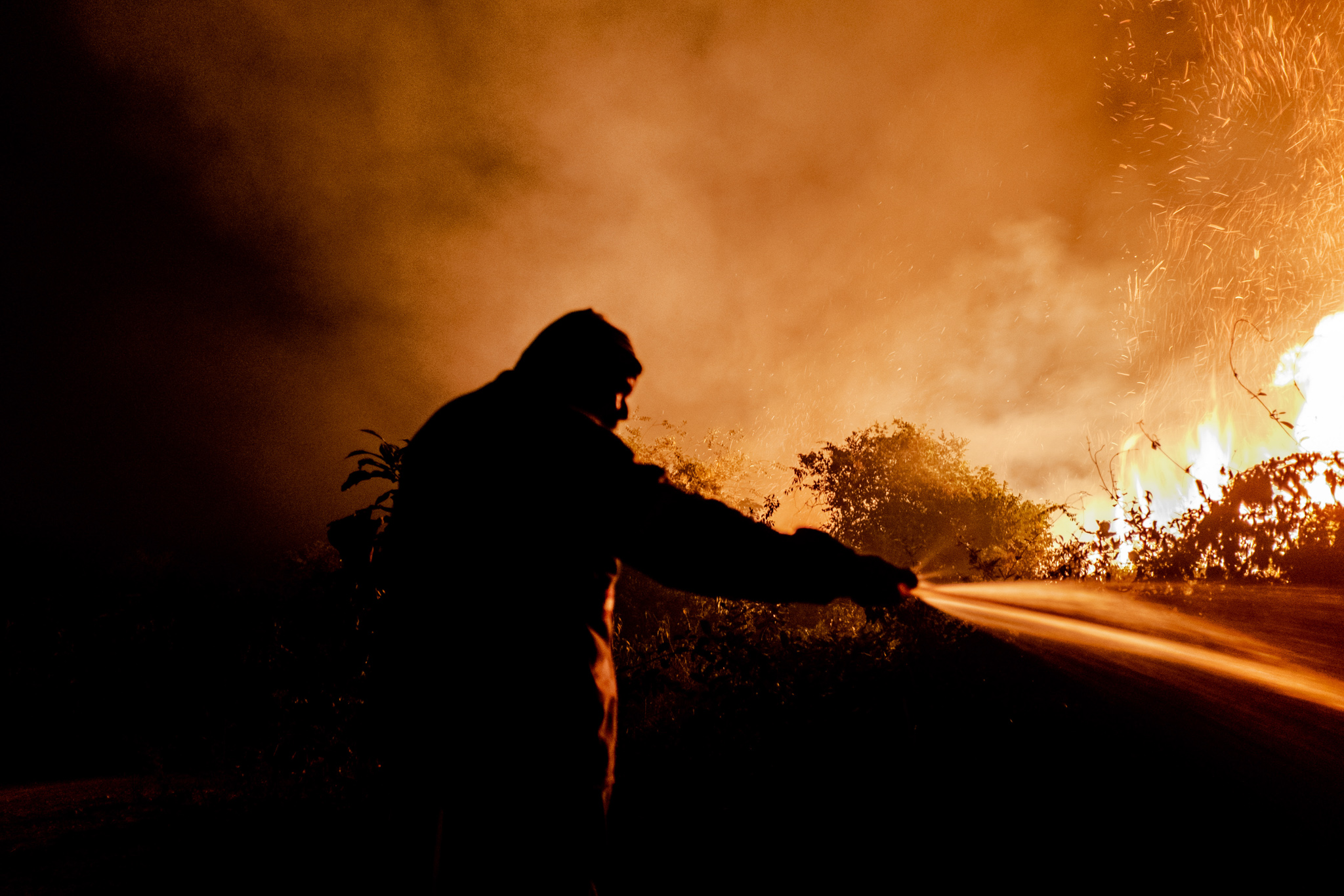 A firefighter sprays water on a blaze, silhouetted against a background of orange smoke