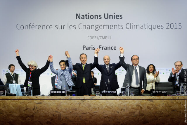 World leaders standing in a line and cheering in front of a United Nations conference backdrop during negotiations for the Paris Climate Agreement