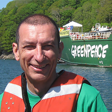 Richard Lancaster, wearing a life jacket, smiles into the camera with a Greenpeace ship visible in the background.