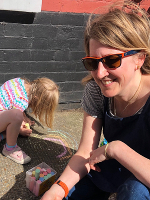 Zoe Hudson in sunglasses smiling up into the camera. A child behind her in drawing on the pavement in colourful chalk