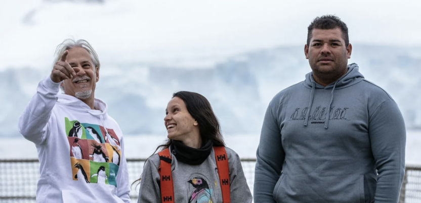 Three Greenpeace crew members wearing branded sweaters with penguin designs. They stand on the deck of a ship in a relaxed, informal pose, with a glacier in the background.