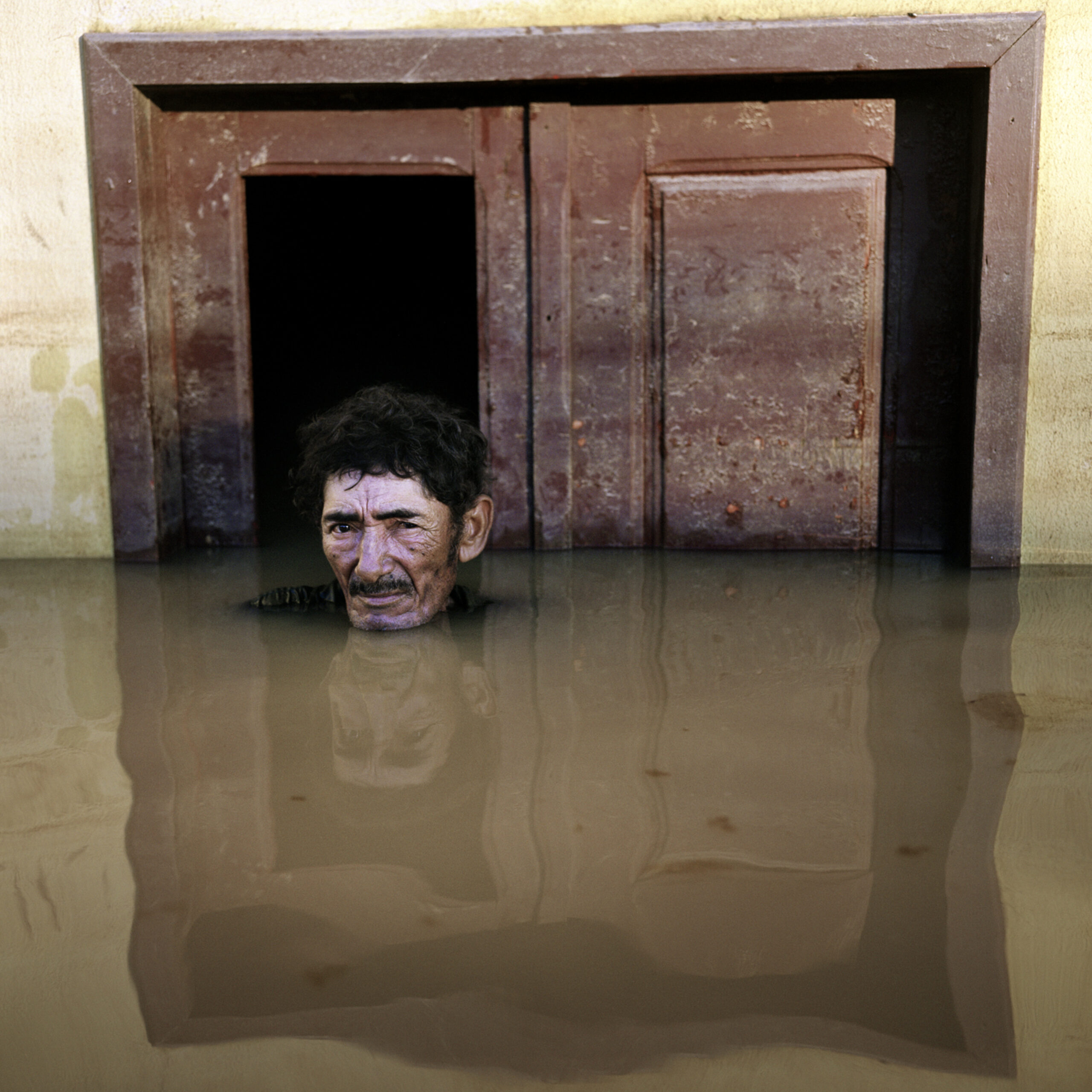 A man is submerged to his shoulders in muddy water while on a street outside a brown door.