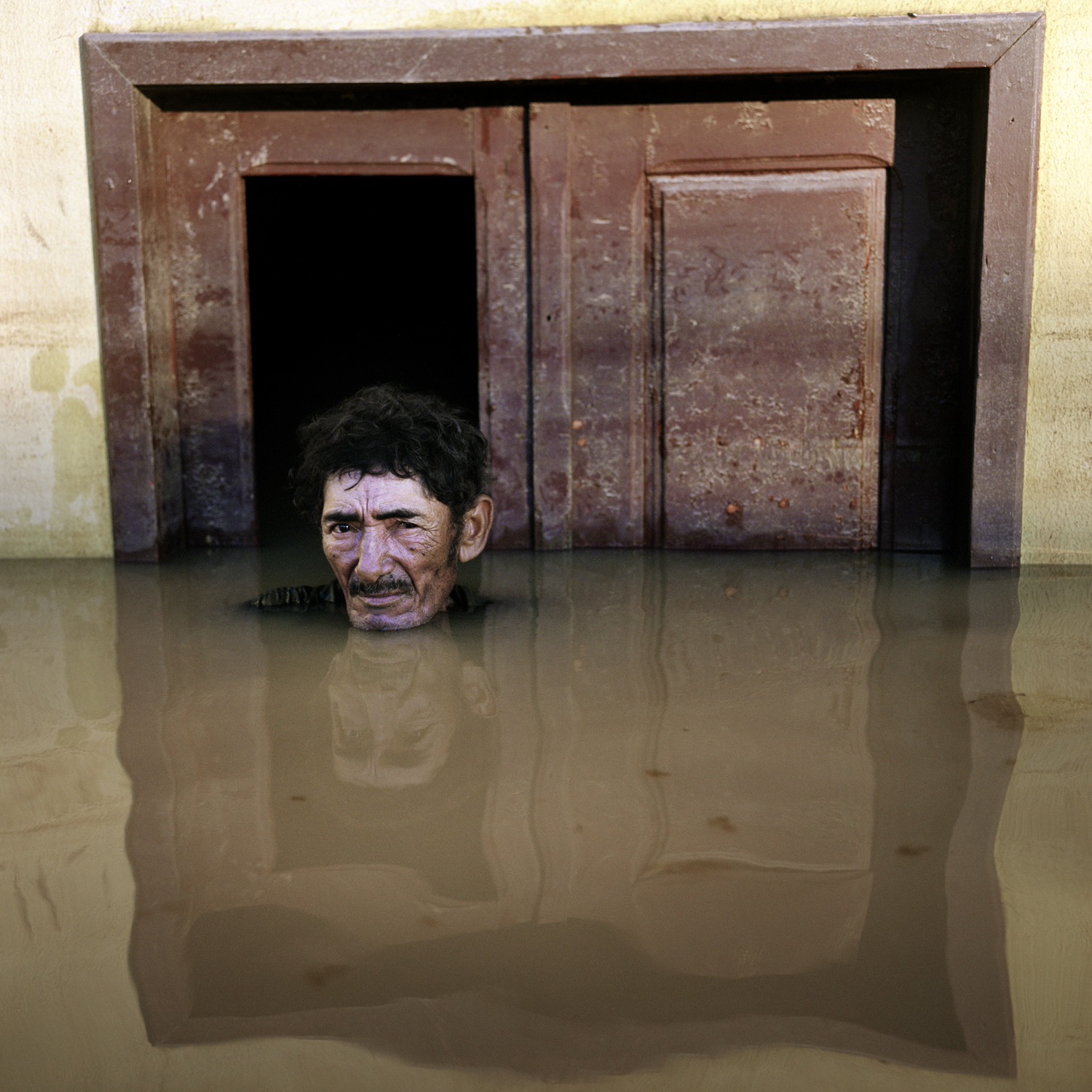A man is submerged to his shoulders in muddy water while on a street outside a brown door.