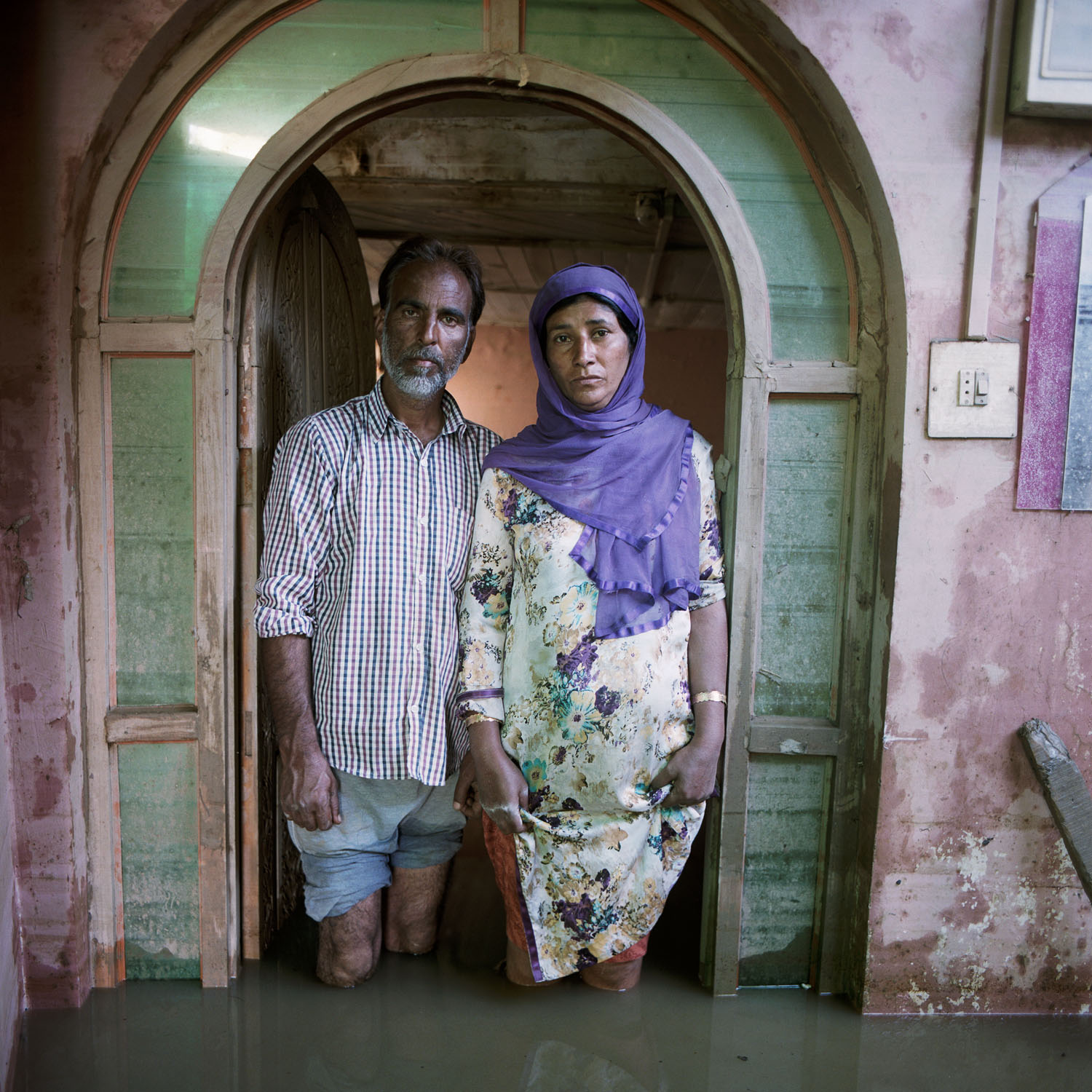 A man and woman stand in a pink and green doorway, with water up to their knees