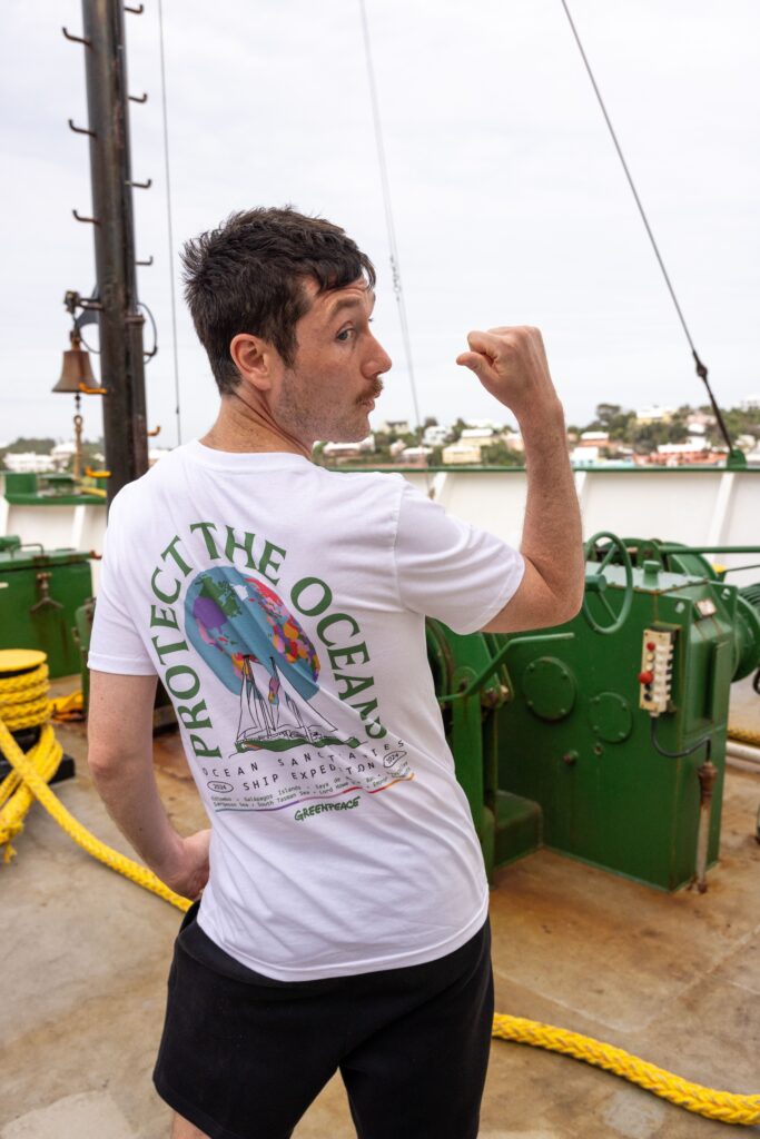 A man with a moustache showing off the back of his T-shirt, while standing on a boat