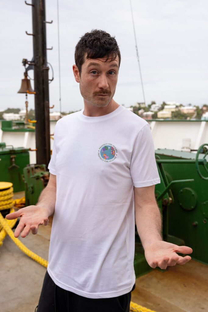 A man in a crisp white T-shirt with a motif on the left chest area stands on a boat
