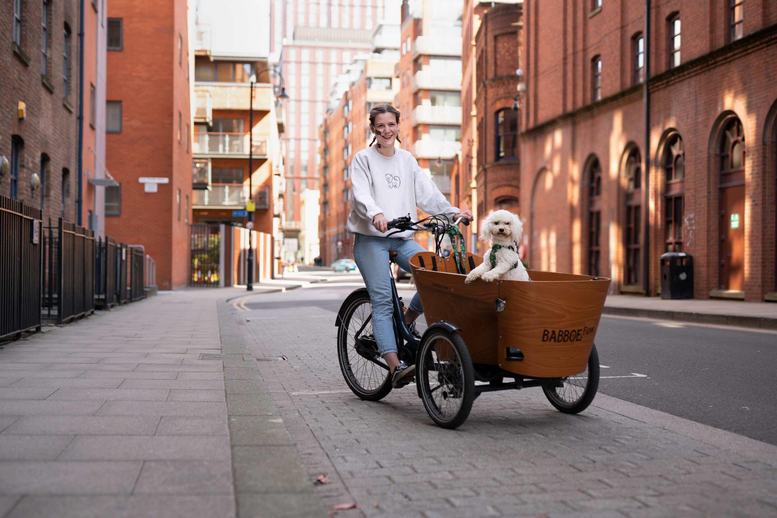 Smiling in the sunshine, Harrie Lannington Spencer rides her cargo trike along a city street. Her small white dog rides in the cargo area up front.