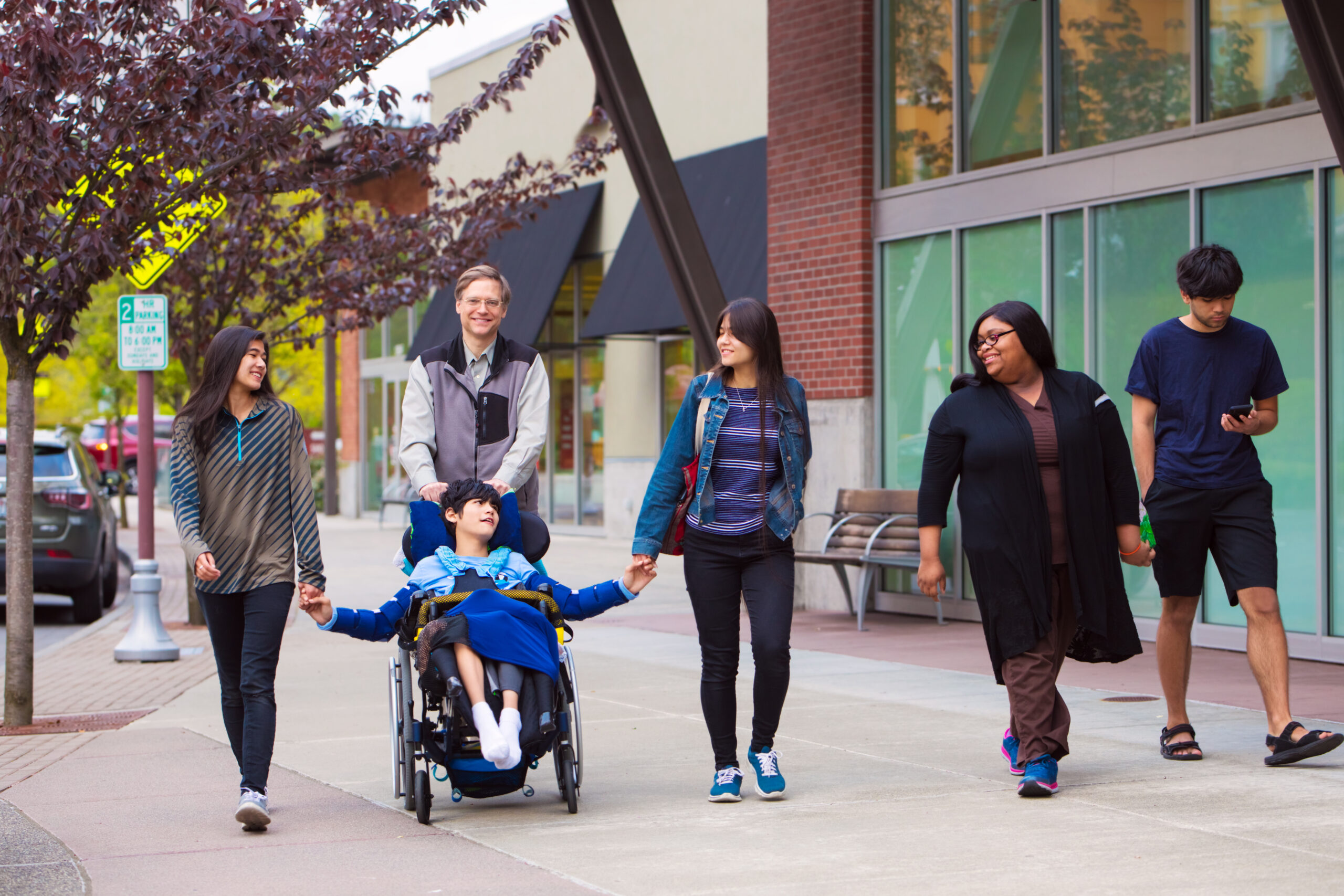 Boy in a wheelchair wheeling on a wide, smooth pavement with a carer and family.