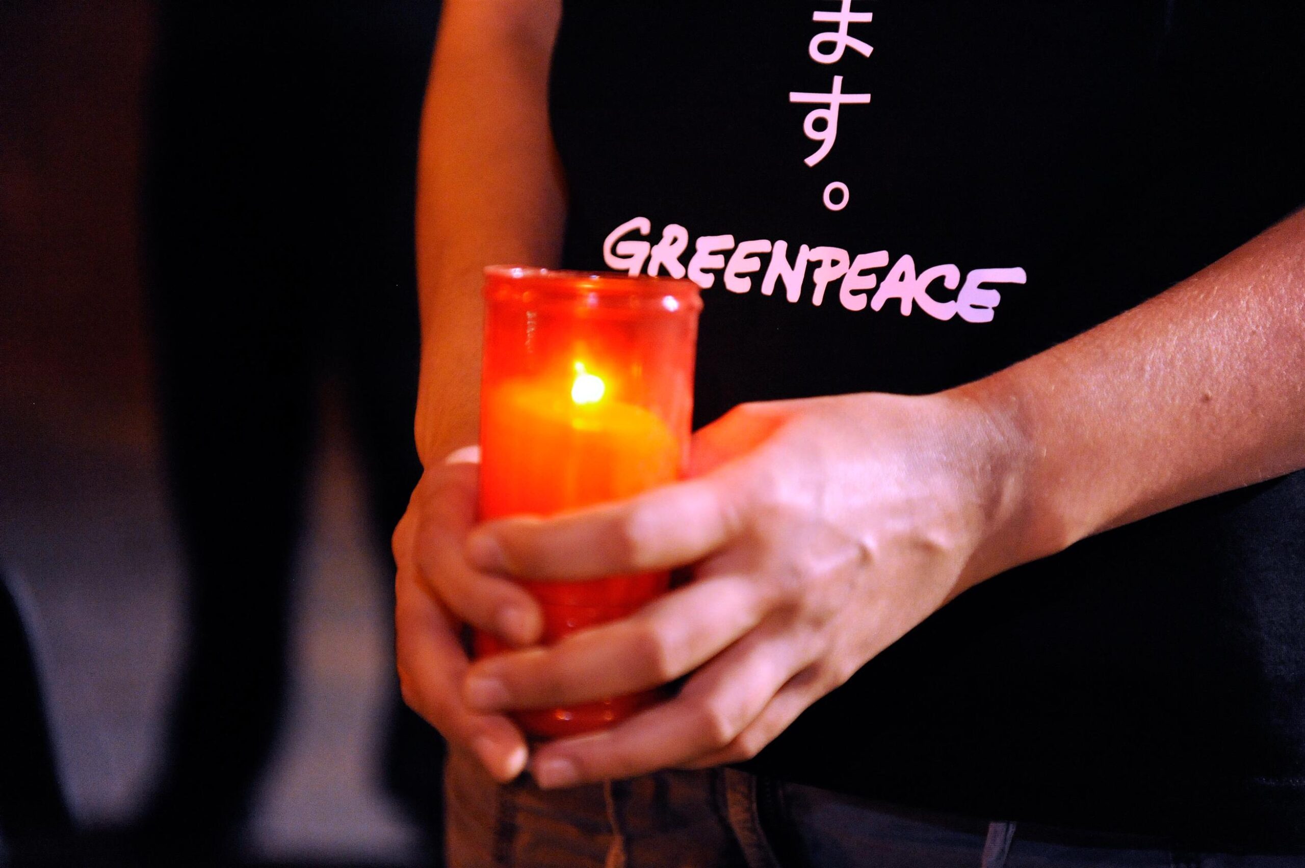 Hands holding a candle in a jar. The holder is wearing a black Greenpeace t-shirt.