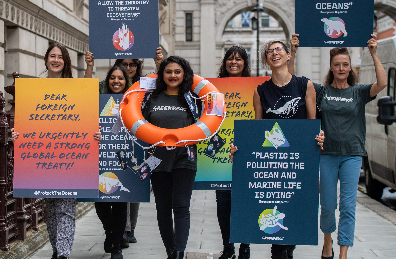 A group of Greenpeace activists walk towards the camera holding up colourful placards calling for action on ocean protection. They're smiling and laughing.