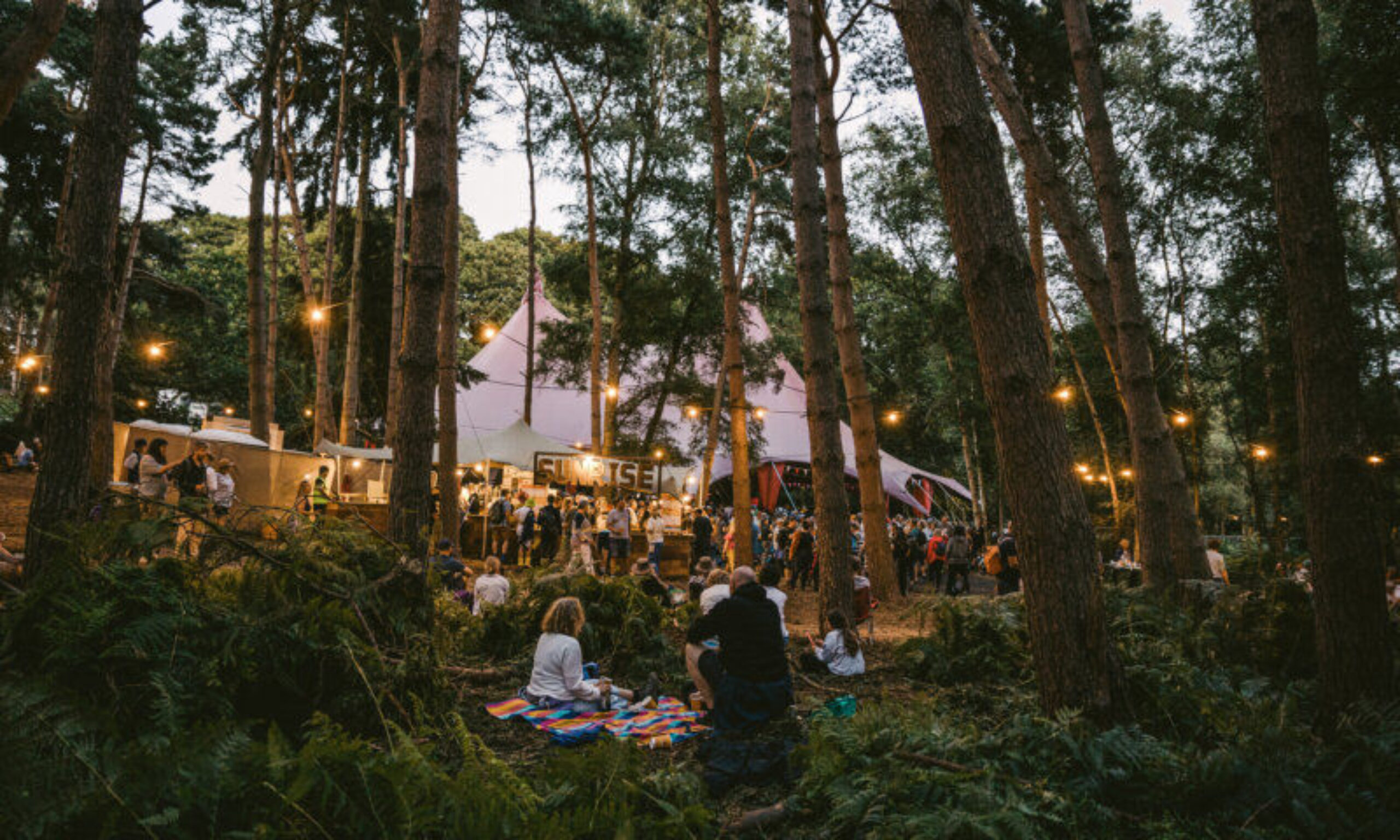 Festivalgoers sit in a lush forest at dusk. Lights are strung through the trees. In the background, a crowd spills out of a marquee tent with an ornate 'Sunrise' sign on the front