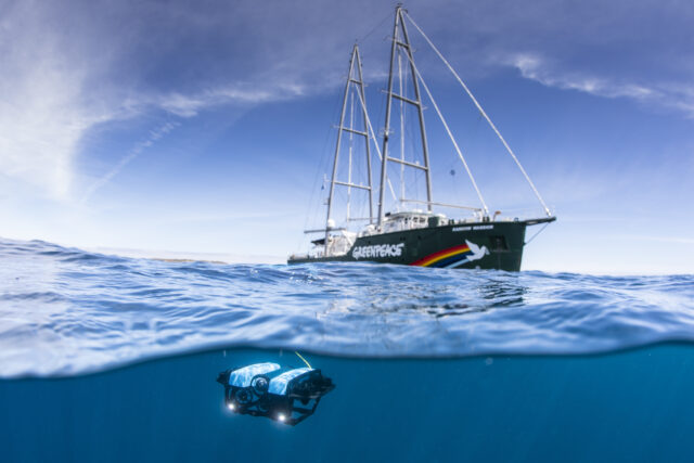A beautiful greenpeace branded ship on a calm sea. The camera lens is half-underwater and a small submersible is visible beneath the ship.