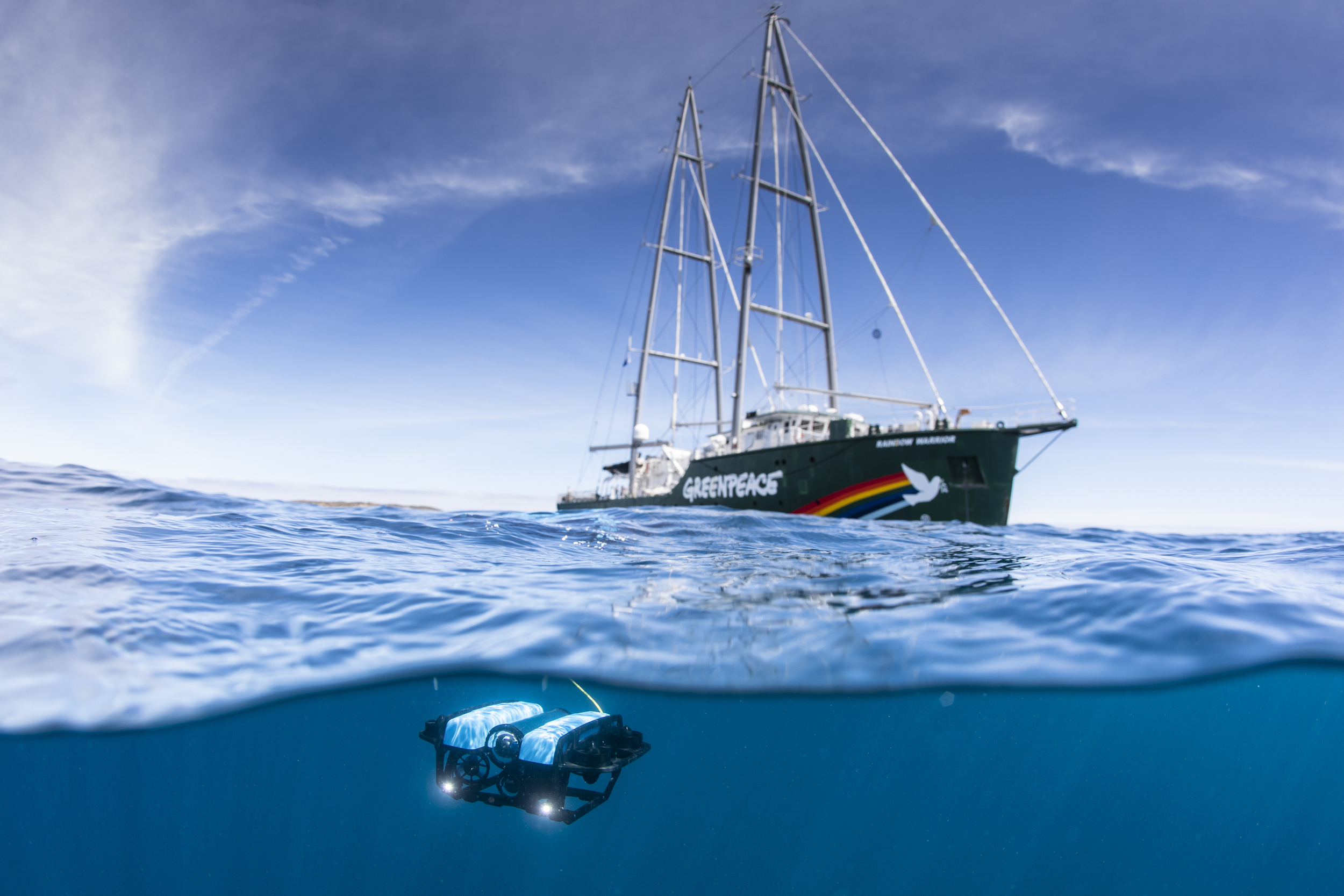 A beautiful greenpeace branded ship on a calm sea. The camera lens is half-underwater and a small submersible is visible beneath the ship.