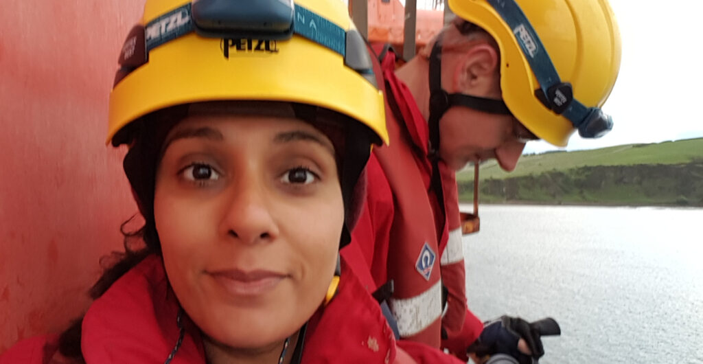 Meena on the oil rig with a yellow helmet and headtorch with Andrew behind her