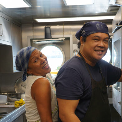 Two members of Greenpeace ship crew smile into the camera while working in the on-board kitchen