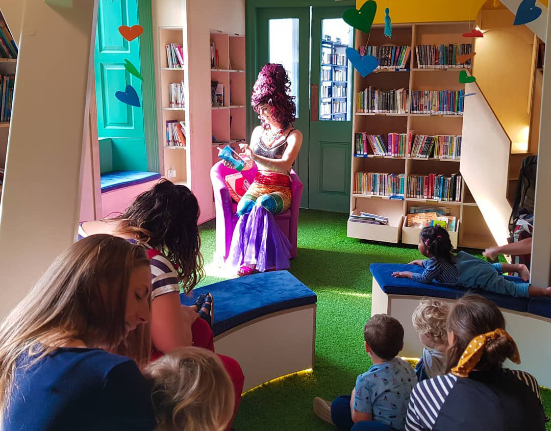 A drag queen reads to a group of children in a colourful sunlit classroom