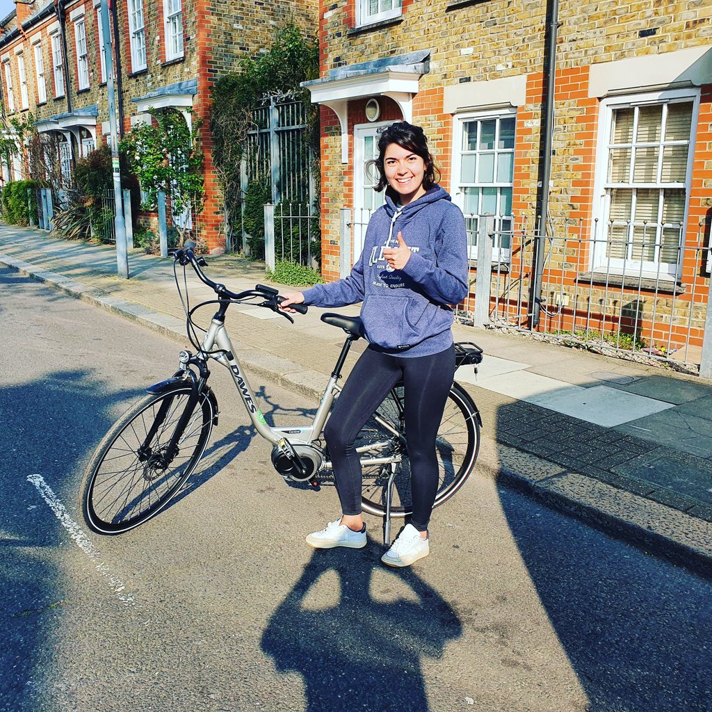 A woman stands in the street with an electric bike, smiling at the camera and giving a thumbs-up