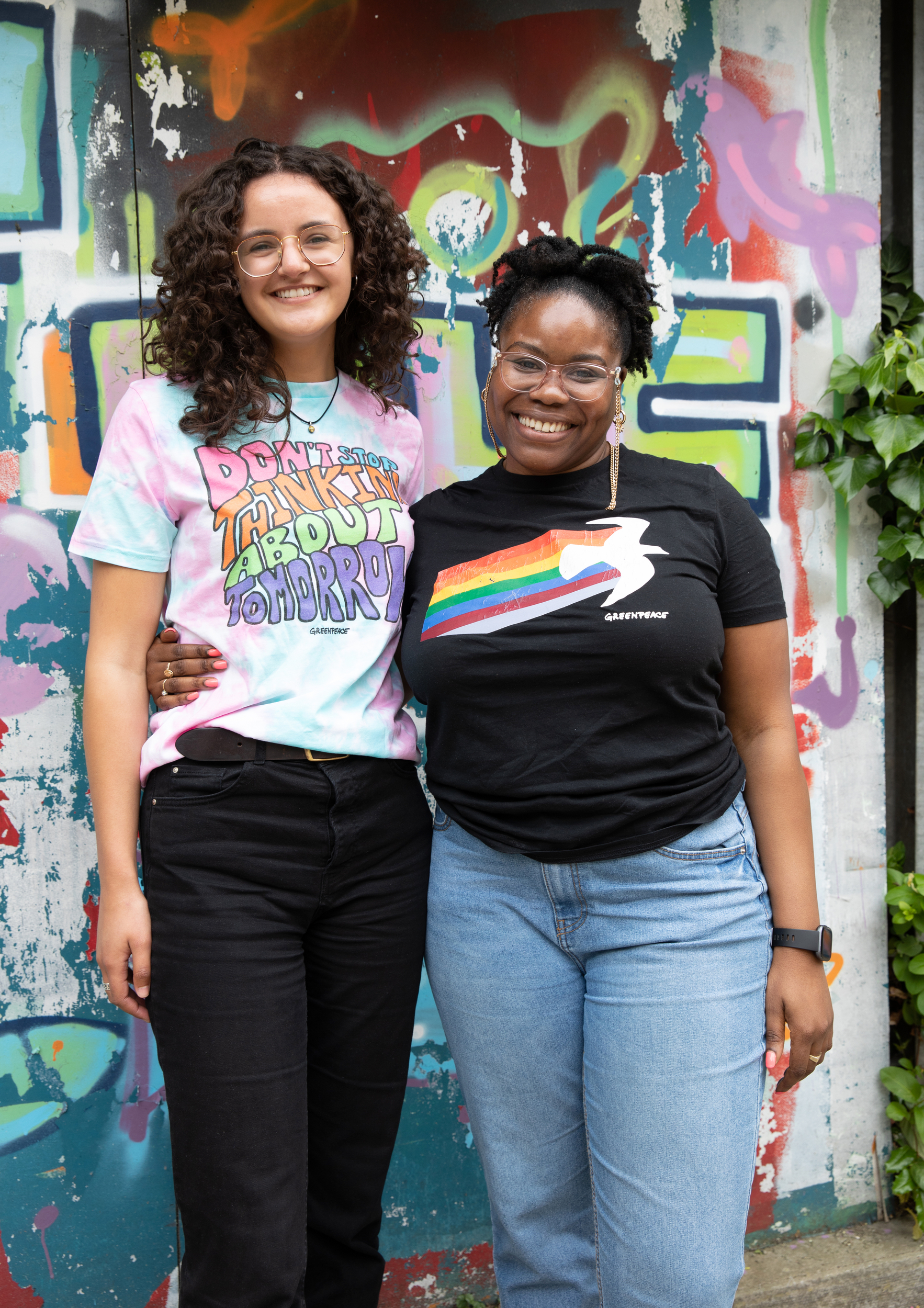 Two women in colourful greenpeace t-shirts hug and smile into the camera