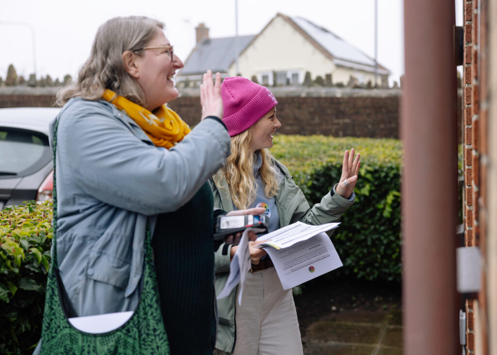 Two voluteers on a doorstep wave to a person off-camera