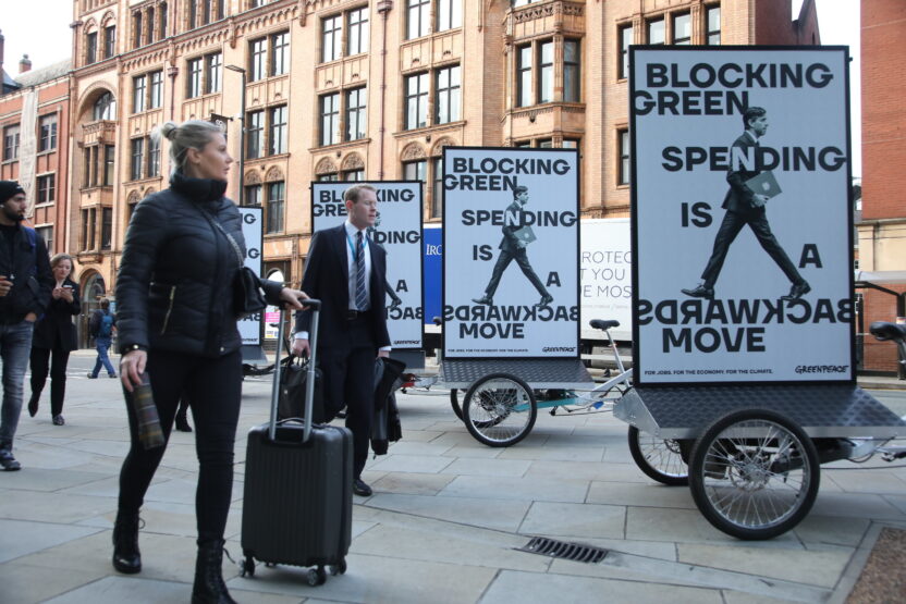 Conference delegates in business suits walk past a trio of cargo bikes with billboards mounted on the back. Each one shows a photo of Rishi Sunak photoshopped so his legs are facing backwards. Text reads 'Blocking green spending is a backwards move'.