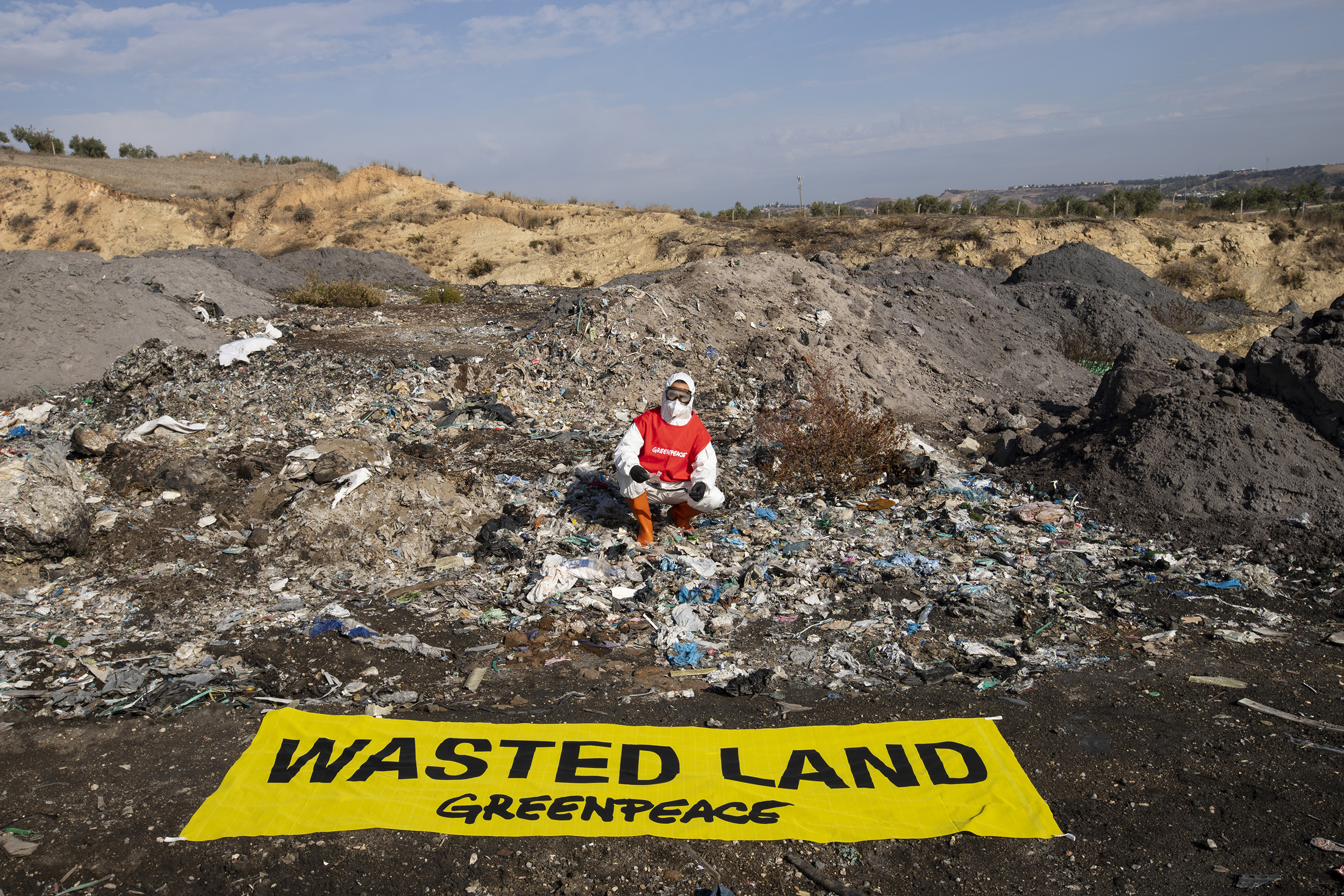 An activist in protective overalls, goggles and a mask kneels in front of a large pile of ash and burned plastic. Laid out in the foreground is a large banner reading 'Wasted land'