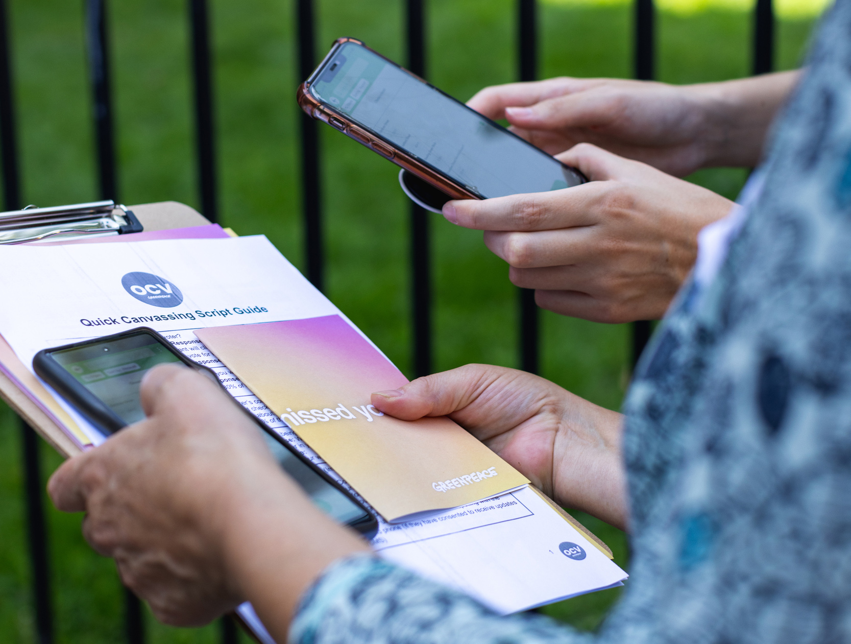 Closeup of two volunteers' hands holding smartphones and a clipboard with paper forms and colourful flyers.