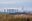 A distant view of several large coal power station chimneys with clouds steaming out of them, and fields in the foreground