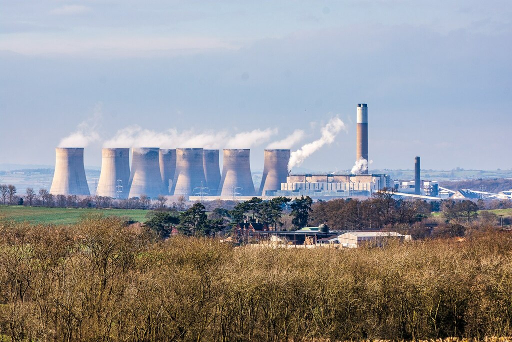 A distant view of several large coal power station chimneys with clouds steaming out of them, and fields in the foreground