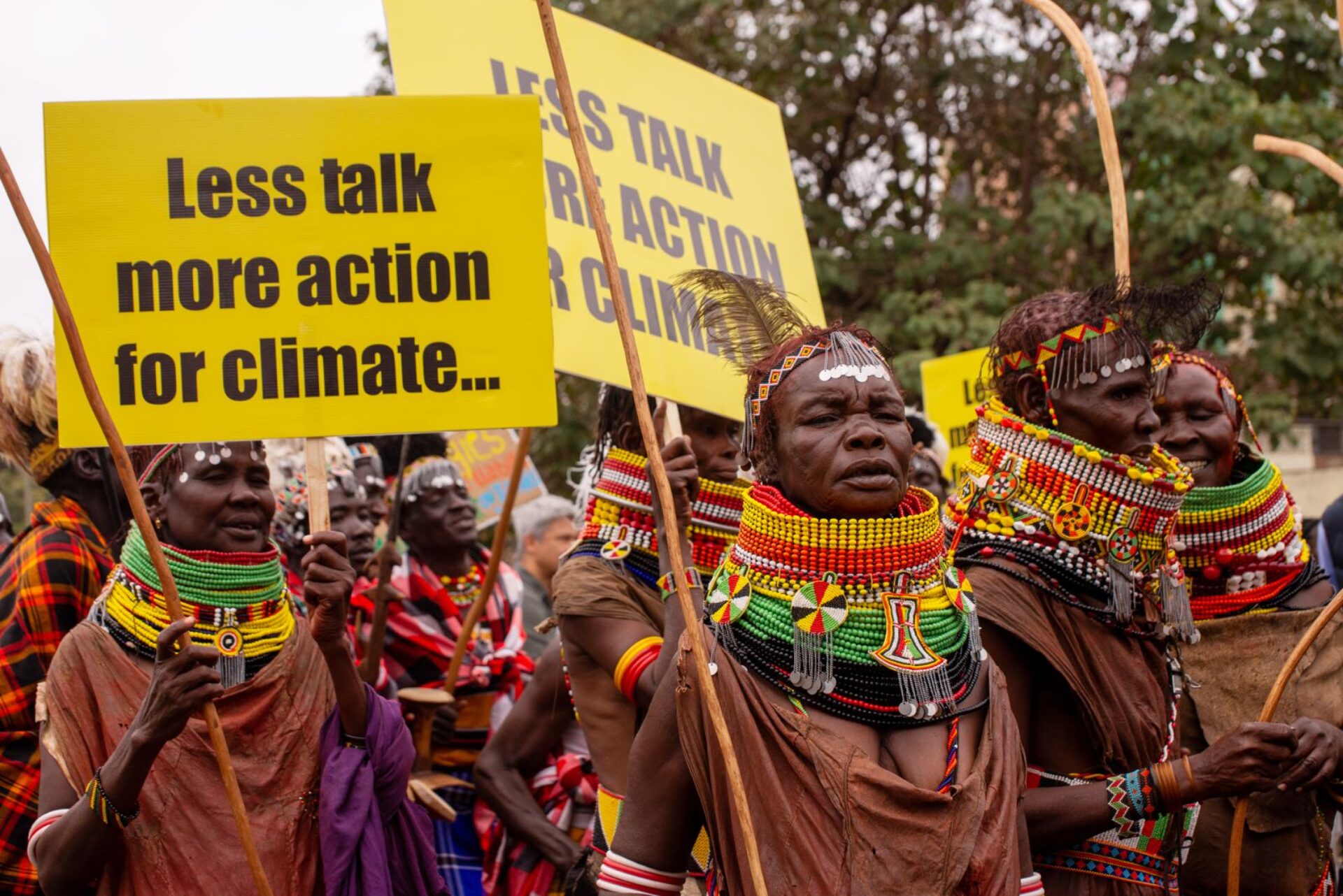 A crowd of Indigenous people with extraordinary large beaded necklace-collars in red, yellow, green and black, with various badges and symbols handing off them. They are holding yellow signs reading Less talk more action for climate...
