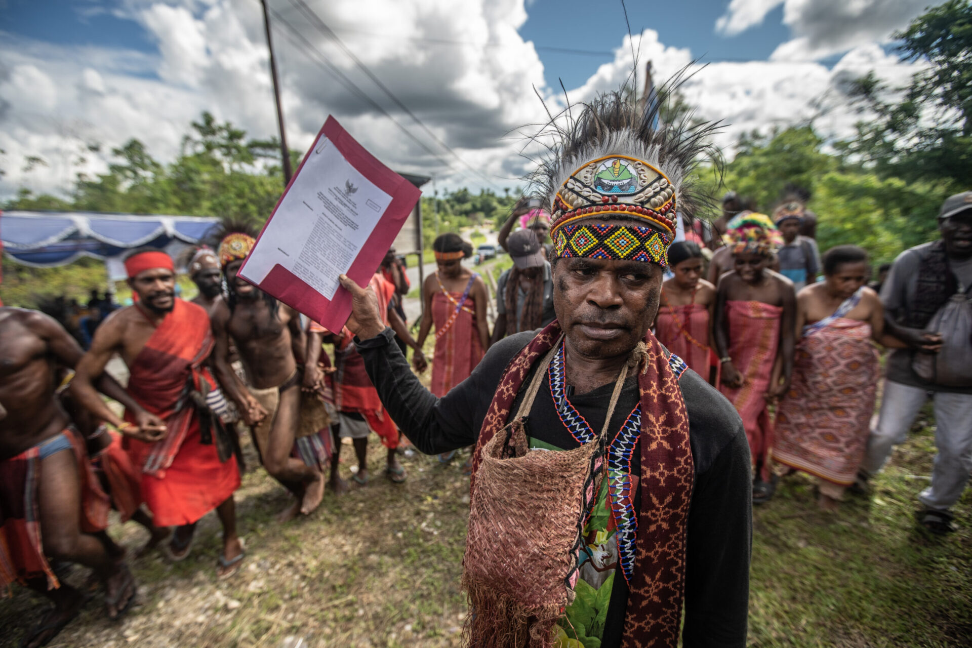A man in feathered and beaded headdress standing in front of a crowd holding up a document in a red folder
