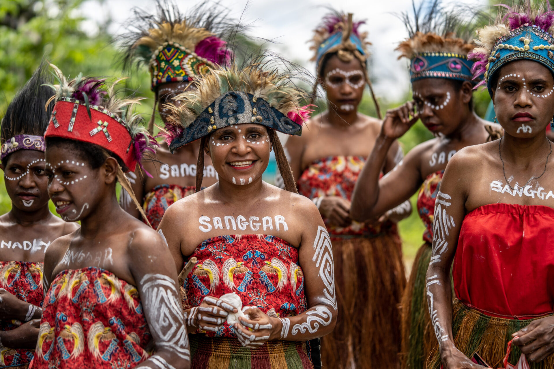 A crowd of women with feather headdresses, face and body paint and colourful clothing. One lady in the middle is smiling brightly at the camera.
